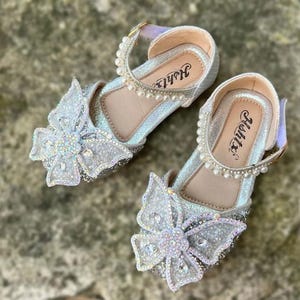 May include: A pair of silver glittery shoes featuring a large butterfly embellishment on the toe, adorned with iridescent rhinestones and a central flower. The ankle strap is decorated with a row of white faux pearls, and the insole displays the text Hshtx.