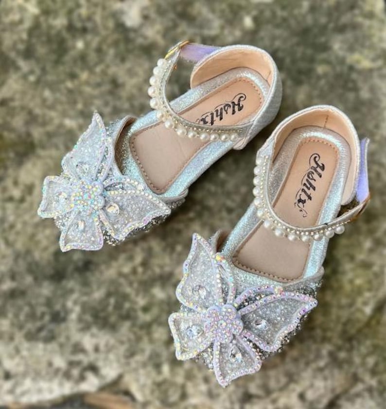 May include: A pair of silver glittery shoes featuring a large butterfly embellishment on the toe, adorned with iridescent rhinestones and a central flower. The ankle strap is decorated with a row of white faux pearls, and the insole displays the text Hshtx.