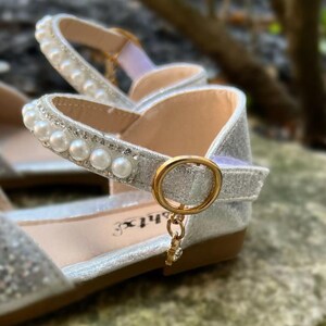 May include: A silver glitter flat shoe featuring an ankle strap adorned with a row of white faux pearls and clear rhinestones. The strap secures with a gold-toned circular buckle and a small dangling charm. The interior sole displays the text "shtx".