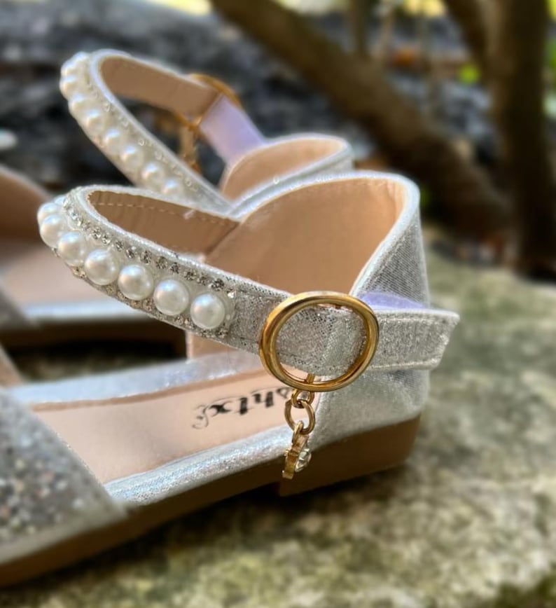 May include: A silver glitter flat shoe featuring an ankle strap adorned with a row of white faux pearls and clear rhinestones. The strap secures with a gold-toned circular buckle and a small dangling charm. The interior sole displays the text "shtx".