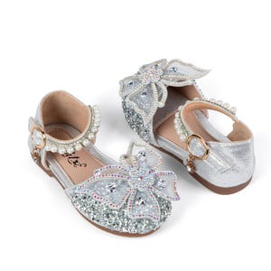 May include: A pair of silver glitter shoes featuring a large decorative bow on the toe adorned with clear crystals and beads. The ankle strap is embellished with a row of white pearls and secures with a gold-tone buckle. The interior is tan.