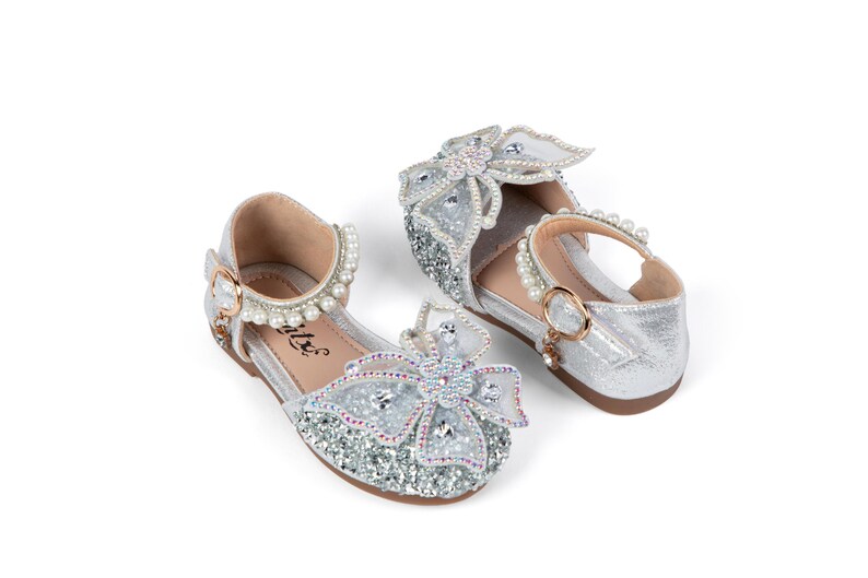 May include: A pair of silver glitter shoes featuring a large decorative bow on the toe adorned with clear crystals and beads. The ankle strap is embellished with a row of white pearls and secures with a gold-tone buckle. The interior is tan.