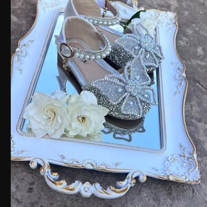 May include: A pair of silver glittery shoes featuring a large butterfly-shaped embellishment covered in clear crystals on the toe, an ankle strap adorned with white faux pearls, and a gold-toned buckle, displayed on a white and gold ornate tray.