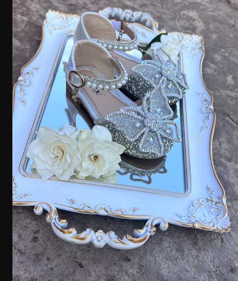 May include: A pair of silver glittery shoes featuring a large butterfly-shaped embellishment covered in clear crystals on the toe, an ankle strap adorned with white faux pearls, and a gold-toned buckle, displayed on a white and gold ornate tray.