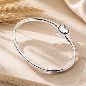 May include: A polished silver bangle bracelet featuring a heart-shaped clasp. The clasp is engraved with the word PANDORA in a circular pattern. The bracelet has a smooth, rounded band design that connects to the heart-shaped closure mechanism.