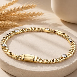 Può includere: Un bracciale a maglie dorate con una chiusura rettangolare incisa con la parola PANDORA. Il bracciale è impreziosito da diverse piccole pietre trasparenti a forma di cuore, posizionate a intervalli lungo le maglie in metallo lucido.