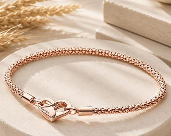 May include: A rose gold-toned bracelet featuring a textured, beaded chain and a heart-shaped clasp. The clasp is engraved with the word PANDORA. The bracelet is circular in shape and has a polished finish on the metal components of the clasp.