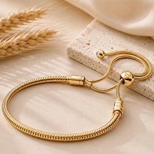 Könnte beinhalten: Ein goldfarbenes Schlangenkettarmband mit einem verschiebbaren, verstellbaren Verschlussmechanismus. Das Armband enthält eine runde Perle mit Gravurdetails und einen kleinen, klaren, runden Stein am Ende der Kette, was ein flexibles Design ergibt.