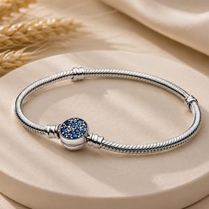 May include: A silver snake chain bracelet featuring a circular clasp. The clasp is decorated with a cluster of small, round, deep blue stones set against a dark background. The bracelet has a smooth, flexible, and segmented metallic texture.