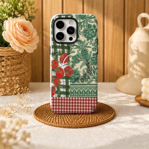 May include: A smartphone case featuring a collage design with red tomatoes on a green plaid background, a green botanical print with leaves and pineapples, and a red and white gingham pattern at the bottom. The case has a circular grip attachment.