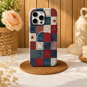 May include: A smartphone case featuring a patchwork design with red, white, and blue squares. The pattern includes various motifs such as stars, stripes, and small text blocks. A circular grip is attached to the centre of the back of the case.