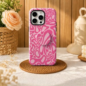May include: A smartphone case featuring a vibrant pink and light pink abstract wavy pattern. The case includes a matching circular collapsible grip mounted on the back, with a cutout for the triple-lens camera system on the upper left corner.