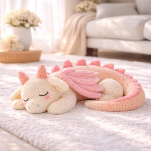 May include: A cream-colored crochet dragon plush toy with pink accents on its horns, wings, and tail spikes. The dragon is in a sleeping position with closed eyes, pink cheeks, and a long, curved tail that wraps around its body on a white surface.