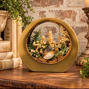May include: A circular gold-toned metal frame containing a decorative arrangement of green fern leaves, small cream-colored berries, and a small brown and white bird figurine resting on a bed of moss within the center opening.