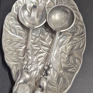 May include: A silver-toned metal serving set consisting of an oval tray with a textured leaf pattern and two matching salad utensils. The handles of the fork and spoon feature sculpted fruit and berry motifs, providing a decorative finish.