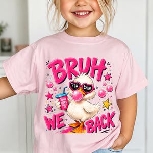 Peut inclure: Un t-shirt rose clair avec un motif représentant un canard blanc portant des lunettes de soleil en forme de cœur, faisant une bulle rose et tenant un gobelet bleu marqué Good Vibes Only. Le texte BRUH WE BACK est écrit en grandes lettres roses.