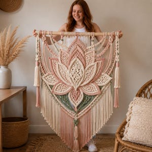 May include: A large macrame wall hanging featuring a central lotus flower design in shades of cream, dusty pink, and sage green. The piece is suspended from a wooden dowel and includes intricate knotted patterns, long fringe, and decorative tassels.