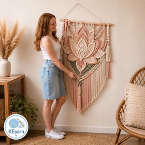 May include: A large macrame wall hanging featuring a central lotus flower design in shades of cream, dusty rose, and sage green. The piece is suspended from a wooden dowel with intricate knotted patterns, long fringe, and decorative tassels.