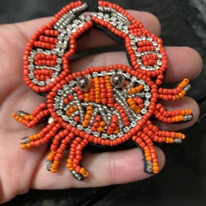 May include: A crab-shaped patch crafted with orange and red seed beads, accented with silver metallic beads and two round silver-toned eyes. The design features a detailed shell and claw structure, with the beads arranged in rows on a black backing.