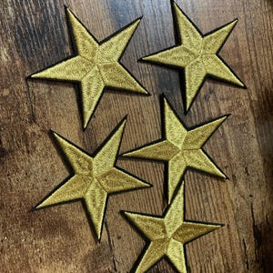 May include: Five embroidered star patches in a metallic gold thread with a black stitched border. Each star features a five-pointed design with detailed thread work radiating from the centre to the points, creating a textured appearance.