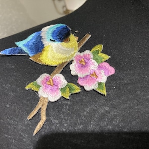 May include: An embroidered patch featuring a small bird with blue, yellow, and white feathers perched on a brown branch. The branch is decorated with several pink flowers with white edges and green leaves, creating a detailed nature-themed design.