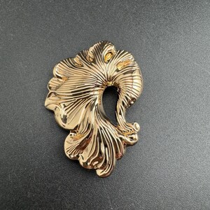 May include: A polished gold-tone metal brooch featuring an abstract, organic shape with deep, flowing ridges and a central open void. The surface is highly reflective, highlighting the curved, fan-like contours of the metallic design.