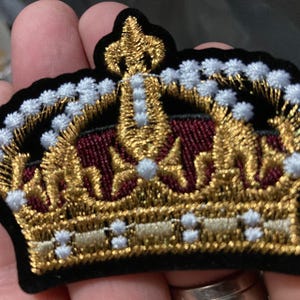 May include: An embroidered patch in the shape of a crown featuring metallic gold thread, a deep burgundy centre, and white accents. The design is outlined in black thread and includes a fleur-de-lis detail at the top centre of the crown structure.