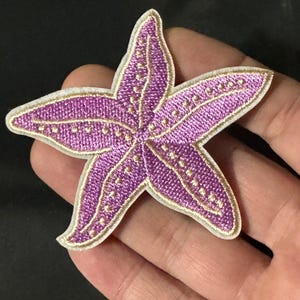 May include: A five-pointed starfish patch featuring vibrant purple embroidery with gold-coloured thread accents along the centre of each arm and a white border edge. The design displays a textured, stitched surface with a distinct star shape.