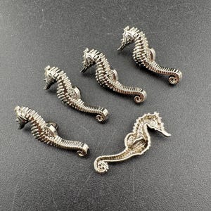 May include: Four silver-toned seahorse-shaped buttons with detailed textured bodies and curved tails. Three of the buttons are oriented with their heads facing upward, while one button is flipped to show its back with a metal shank attachment.