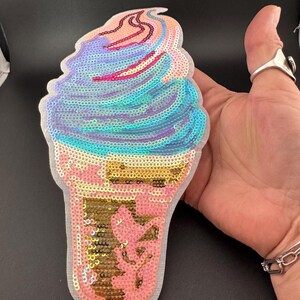 May include: An ice cream cone patch covered in iridescent sequins. The top features swirls of blue, purple, and pink, while the cone section is rendered in gold and pink sequins. The entire shape is outlined with a white fabric border.