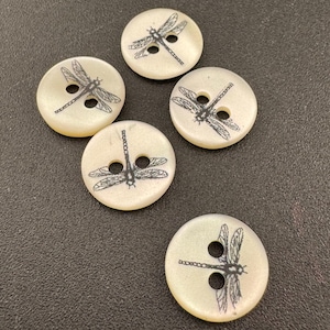 May include: Five round, off-white buttons, each featuring a black printed dragonfly graphic. Every button has two central holes for attachment. The surface of the buttons has a smooth, slightly pearlescent finish, and the dragonfly designs are oriented in various directions across the set.