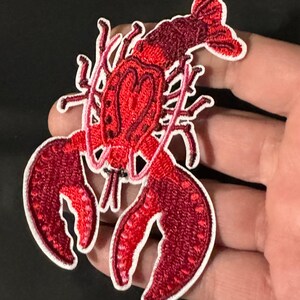 May include: An embroidered lobster patch featuring vibrant shades of red and dark crimson thread with a white border. The design includes detailed stitching for the claws, body, and tail segments, presented against a plain background.