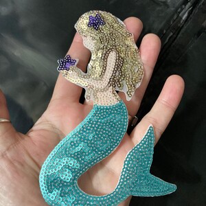 May include: A mermaid-shaped appliqué patch covered in sequins. The mermaid has gold-coloured hair, a teal-coloured tail, and is holding a small purple star-shaped sequin. A second purple star-shaped sequin is positioned in the mermaid's hair.
