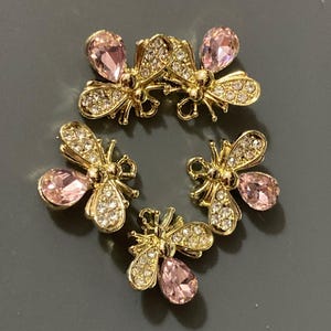 May include: Four bee-shaped ornaments crafted from gold-toned metal. Each bee features a large, pear-shaped light pink crystal on the abdomen and smaller, clear round rhinestones embedded along the wings and body, creating a sparkling effect.