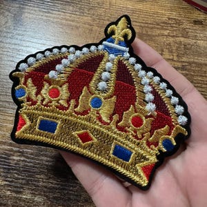 May include: An embroidered patch shaped like a crown with a gold base, red fabric panels, and a fleur-de-lis at the top. The design features blue and red square accents, white beaded-style embroidery along the arches, and a black border.