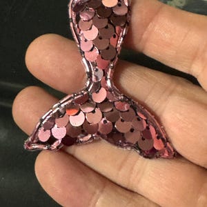 May include: A mermaid tail patch covered in shimmering pink sequins. The decorative applique features a distinct fin shape with a stitched border, showcasing a reflective surface that catches the light with various shades of pink and rose tones.