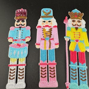 May include: Three embroidered nutcracker patches featuring sequin embellishments. The left figure wears a pink hat and blue jacket with a drum. The centre figure wears a blue hat and pink jacket. The right figure wears a gold crown and yellow jacket.