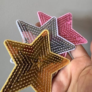 May include: Three star-shaped patches featuring dense sequin embellishments. The front patch is gold, the middle patch is silver with a white border, and the back patch is pink. Each star has a distinct felt-like backing visible along the edges.