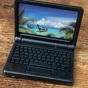 May include: A miniature black laptop toy with a screen displaying a tropical beach scene featuring two traditional long-tail boats on turquoise water, surrounded by palm trees under a bright blue sky with white clouds, and a black keyboard base.
