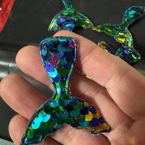 May include: A set of three mermaid tail-shaped patches covered in iridescent sequins in shades of blue, green, and purple. The patches are approximately 7.6 cm tall, as indicated by the visible portion of a ruler in the bottom left corner.