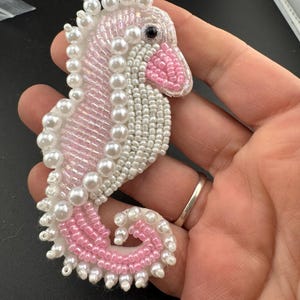 May include: A seahorse-shaped brooch crafted with intricate beadwork and pearls. The design features a combination of iridescent white, soft pink, and clear glass seed beads, accented with round white pearls along the spine and a black bead eye.