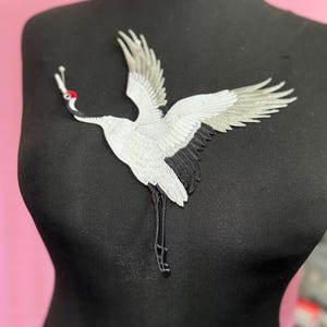 May include: An embroidered patch featuring a crane in flight with white and grey wings, a black body, and a distinct red crown on its head. The bird is depicted with its wings spread wide, a long neck, and thin black legs extending downwards.