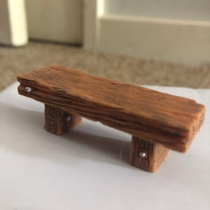 May include: A miniature bench crafted from brown material with a textured wood grain finish. The seat is a single thick plank supported by two block-style legs. Small, circular silver-coloured metal accents are embedded into the sides of the legs.