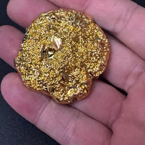 May include: A circular, metallic gold-coloured object with a highly textured, uneven, and reflective surface. The piece features a dense, granular, and crinkled appearance with small, smooth, and shiny highlights scattered across its irregular form.