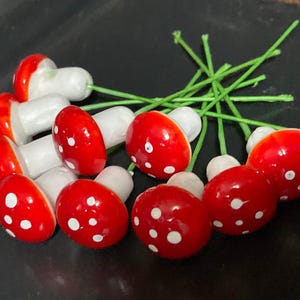 May include: A collection of small, decorative mushroom ornaments featuring glossy red caps with white spots and white stems. Each mushroom is attached to a thin, flexible green wire stem, allowing for easy placement in various craft projects.