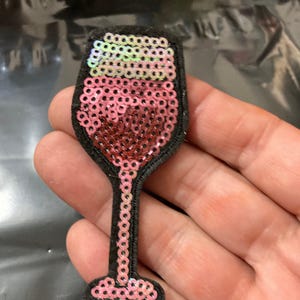 May include: A wine glass shaped patch featuring iridescent pink and red sequins on a black felt backing. The design includes a stem and base outlined in pink sequins, with the bowl filled with red sequins and a top layer of iridescent sequins.