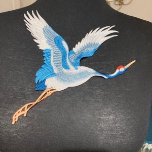May include: An embroidered patch featuring a crane in flight with white and blue wings, a long neck, a red patch on its head, and light orange legs. The bird is depicted in a dynamic pose with wings spread wide, showcasing detailed stitching.