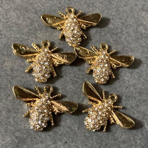 May include: Five gold-toned bee charms arranged in a group. Each bee features a body encrusted with clear, sparkling rhinestones and smooth, polished gold-coloured wings. A small circular loop is attached to the top of each bee for hanging.