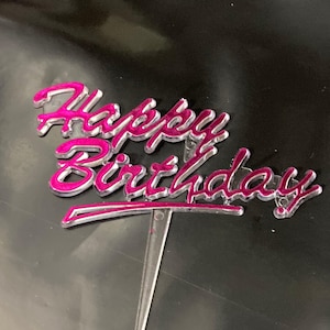 May include: A cake topper featuring the text Happy Birthday in a cursive font. The lettering is constructed from clear acrylic with a vibrant hot pink inlay. A single clear acrylic stake extends downward from the centre of the design.