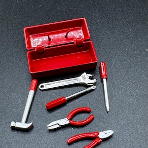 May include: A miniature red metal toolbox with an open lid, accompanied by a set of small tools including a silver hammer with a red handle, a silver adjustable wrench, a screwdriver with a red handle, and two pairs of pliers with red handles.